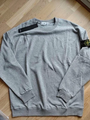 Grå sweatshirt från Stone Island - Klassisk grå sweatshirt från Stone Island med rund hals och ribbade muddar. Tröjan har den ikoniska svarta patchen med gul och grön kompass på vänster ärm. Tillverkad i mjuk bomull för skön passform och stilren look.