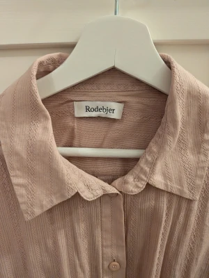 Beige skjortklänning Rodebjer XS - Lång beige skjortklänning från Rodebjer i 100% bomull. Modellen har knappar framtill, lång ärm med manschett och ett snyggt knytband i midjan. Tunt randigt tyg med diskret struktur och klassisk krage. Perfekt för en chill och stilren look. Endast använd en gång!