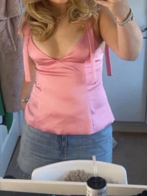 Rosa satinlinnetopp med knytband - Supersöt rosa linnetopp i satin med smala axelband som knyts på axlarna. Toppen har en v-ringning. Perfekt för sommarens alla tillfällen och lätt att styla med kjol eller jeans.