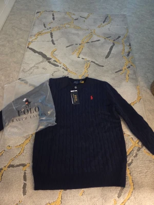 Mörkblå kabelstickad tröja Polo Ralph Lauren - Kabelstickad mörkblå tröja från Polo Ralph Lauren i 100% bomull. Klassisk rund halsringning och röd broderad logga på bröstet. Långärmad modell med ribbade muddar och snygg struktur. Perfekt till jeans eller chinos.
