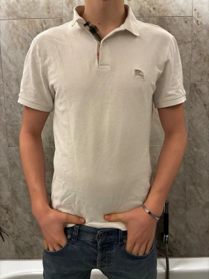 Vit pikétröja från Burberry - Säljer en klassisk vit pikétröja från Burberry med korta ärmar och den ikoniska rutiga detaljen vid knäppningen. Tröjan har en liten broderad Burberry-logga på bröstet och är gjord i mjuk bomull. Perfekt till jeans eller shorts för en clean look.