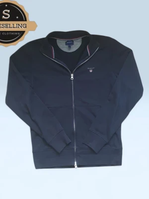 Mörkblå zip-kofta från GANT, S - Snygg mörkblå kofta från GANT med hel dragkedja och broderad logga på bröstet. Ståkrage med diskreta rödvita detaljer på insidan. Tillverkad i mjuk bomull, perfekt för lager-på-lager. Klassisk och stilren design som funkar året runt.