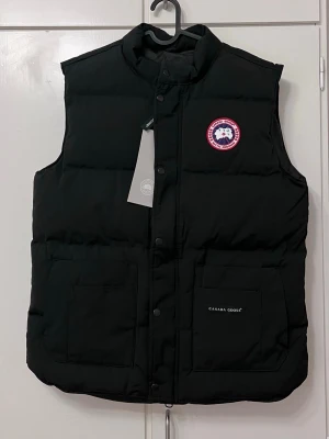 Svart dunväst från Canada Goose L - Säljer en svart dunväst från Canada Goose, modell Freestyle Vest. Västen har hög krage, två stora framfickor och stängs med tryckknappar. Klassisk Canada Goose-logga på bröstet och diskret broderad logga på fickan. Perfekt för lager-på-lager under kyliga dagar.