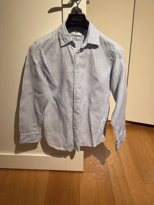 Randig linneskjorta från Uniqlo XS - Fräsch skjorta från Uniqlo i storlek XS, tillverkad i 100% linne. Den har ljusblå och vita vertikala ränder, klassisk krage och knappar framtill. Perfekt för dig som gillar chill och stilren look med långärmad passform.