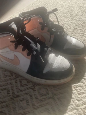 Nike Air Jordan 1 Peach/Black/White - Säljer ett par Nike Air Jordan 1 med snygg färgkombo i persikofärgat, svart och vitt. Skorna har klassisk snörning, hög siluett och den ikoniska swoosh-loggan på sidan. Tillverkade i skinn och syntetmaterial. Perfekta för dig som vill sticka ut med dina sneakers.