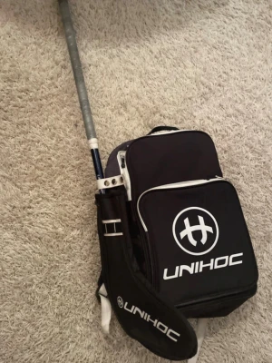 Svart ryggsäck från Unihoc - Säljer en svart ryggsäck från Unihoc väldigt lätt tillgänglig innan träning, med vita detaljer och logga. Väskan har 2 fack med dragkedja och ett specialfack för innebandyklubbor då jag själv får plats med 2 st i samma fack.  Tillverkad i slitstark syntet och har vadderade axelband för extra komfort. Perfekt för träning.