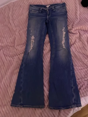 Blå bootcut jeans med slitningar - Säljer ett par blå bootcut jeans med slitna detaljer på låren och snyggt tvättad finish. Jeansen har låg midja, perfekta för dig som gillar en retro och avslappnad stil.