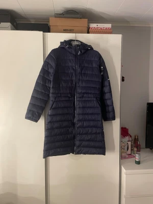 Mörkblå lång ridjacka CRW S - Lång, mörkblå pufferjacka från CRW med huva och dragkedja framtill. Jackan har quiltad design, praktiska fickor med dragkedja och tryckknappar baktill för extra rörelsefrihet. Perfekt för kalla dagar och riktigt snygg till vardagslooken.