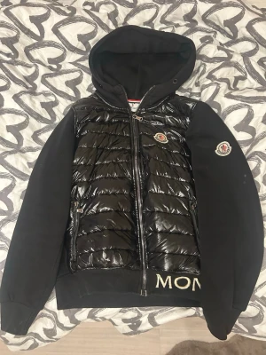Svart pufferjacka från Moncler - Säljer en svart pufferjacka från Moncler med glansig quiltad front och mjuka ärmar i sweatshirtmaterial. Jackan har huva, dragkedja framtill och broderad Moncler-logga på bröstet och ärmen. Ribbad nederkant med broderad text. Snygg och stilren look. Stl S/M