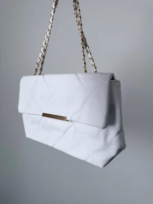 Vit Axelväska - Snygg vit axelväska med quiltat mönster och guldfärgad kedja som axelrem. Väskan har ett stilrent metallspänne framtill och magnetlås. Tillverkad i skinnimitation med mjuk känsla. Perfekt för dig som gillar klassisk och trendig design.