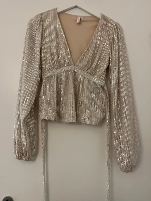 Glittrig topp från Nelly - Säljer en champagnefärgad omlottopp från Nelly med långa puffärmar och djup v-ringning. Toppen är täckt av glittriga paljetter och har knytband i midjan för en snygg siluett. Använd en gång