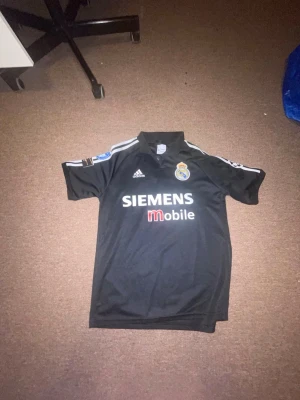 Real Madrid Zidane svart matchtröja - Säljer en svart Real Madrid matchtröja från Adidas med Zidane och nummer 5 på ryggen. Tröjan har vita detaljer, klubbmärke och Siemens Mobile-tryck på bröstet. Perfekt för fotbollsfans som vill sticka ut på planen eller läktaren.