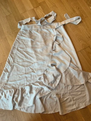 Beige omlottkjol med volang från Chelsea Girls - Supersöt beige omlottkjol från Chelsea Girls med volangdetaljer längs kanten. Kjolen har knytband i midjan och är perfekt för dig som gillar en feminin och trendig stil. Lätt och luftig modell som passar till många olika outfits.