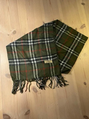 Grön rutig halsduk från Burberry - Stilren halsduk i grönt med klassiskt rutmönster i vitt, svart och rött från Burberry. Halsduken har fransar i ändarna och är tillverkad i mjukt tyg som känns skönt mot huden. Perfekt accessoar för att lyfta din outfit. Pris kan diskuteras.