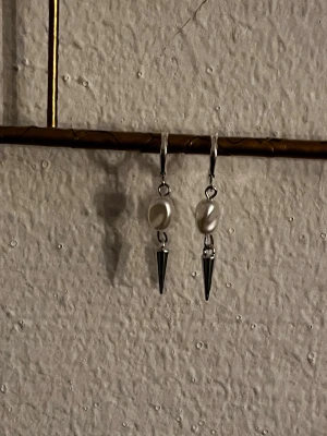Silvriga örhängen med pärla och spike - Snygga örhängen i silverfärg med oval vit pärla och en cool konformad spike. Hängande modell som ger en edgy och trendig look. Perfekta för dig som gillar att sticka ut med dina accessoarer.