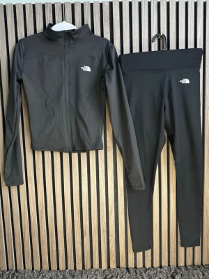 Svart träningsset från The North Face - Snyggt svart träningsset från The North Face med långärmad zip-jacka och matchande tights. Setet är gjort i stretchigt material, 78% polyamid och 22% spandex, vilket ger en skön och följsam passform. Perfekt för gymmet eller löprundan.