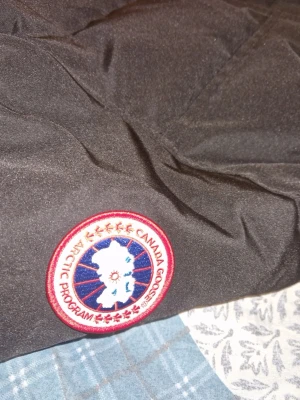 Svart pufferjacka från Canada Goose - Svart pufferjacka från Canada Goose med huva och klassisk logga på ärmen. Jackan har dragkedja och knappar framtill, två stora fickor och quiltad design. Materialet är polyester och dun, perfekt för kalla dagar. Snygg och funktionell för dig som vill ha värme och stil. Jackan släpper ganska mycket fjädrar på insidan och är lite sönder på högersidan.
