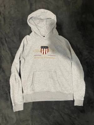 Grå hoodie från GANT med tryck - Snygg grå hoodie från GANT med broderad logga och texten 'American Sportswear' på bröstet. Klassisk känguruficka framtill, ribbade muddar och huva. Perfekt för en avslappnad och sportig stil. Mjuk bomullsblandning för extra komfort.