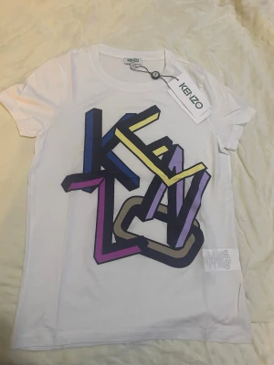 Vit Kenzo t-shirt med färgglatt tryck - Snygg vit t-shirt från Kenzo med ett stort, färgglatt grafiskt tryck framtill i blått, gult, lila och svart. Klassisk passform med rund hals och korta ärmar. Tillverkad i mjuk bomull för en bekväm känsla. Perfekt för dig som vill sticka ut med en trendig look.