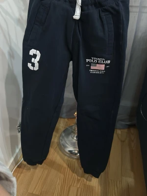 Mörkblå mjukisbyxor Vinson Polo Club - Mörkblå mjukisbyxor från Vinson Polo Club med vita tryckdetaljer, snörning i midjan och två fickor fram. Snygg sportig look med siffran 3 på benet och logga på låret. Perfekta för chill eller träning.