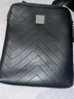 Svart quiltad handväska från BOSS - Snygg svart handväska från BOSS med quiltad design i sicksackmönster. Väskan har en stilren fyrkantig form och ett diskret BOSS-märke framtill. Tillverkad i skinnliknande material och har en bred axelrem för extra komfort.