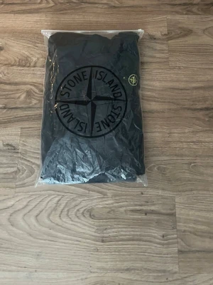 Svart vindjacka från Stone Island - GÅR EJ NER I PRIS.Snygg svart vindjacka från Stone Island med huva och dragkedja framtill. Jackan har den klassiska logotypen på ärmen och är tillverkad i lätt polyester. Perfekt för dig som vill ha en stilren och sportig look.