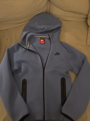 Blå hoodie från Nike med dragkedja - Snygg blå hoodie från Nike med svart logga på bröstet och hel dragkedja framtill. Två svarta fickor med dragkedja och huva. Perfekt för chill dagar eller när du vill ha en sportig look.