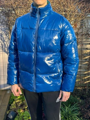 Blå glansig pufferjacka från ASOS - Ovanlig och slutsåld snygg blå pufferjacka från ASOS med glansig finish och hög krage. Jackan är helt ny och oanvänd! Person på bilden är 180 cm, se bilder för mått. Perfekt för kalla dagar när du vill sticka ut med en modern look.