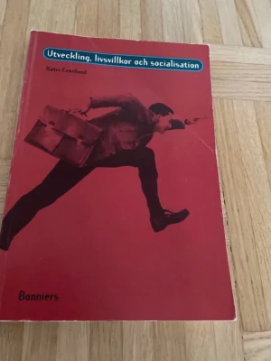 Utveckling, livsvillkor och socialisation - Kursbok med röd framsida och svartvit bild, skriven av Katri Cronlund. Boken tar upp frågor om utveckling, livsvillkor och socialisation, och är anpassad för gymnasiekurser inom psykologi och samhällsämnen.