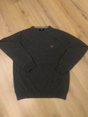 Mörkgrå stickad tröja från Gant - Mörkgrå stickad tröja från Gant i superfin lammull. Klassisk rund halsringning med ribbade muddar och diskret Gant-logga broderad på bröstet. Perfekt för dig som gillar stilrena och tidlösa plagg.  Pris kan diskuteras vid intresse