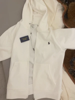 Vit hoodie från Polo Ralph Lauren - Säljer en fräsch vit hoodie från Polo Ralph Lauren med dragkedja framtill och klassisk huva. Hoodien har två fickor och den ikoniska lilla loggan broderad på bröstet. Perfekt för en clean och stilren look.