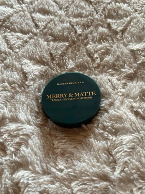 Merry & Matte Setting Powder - Translucent setting powder från Makeup Mekka i en rund, mörkgrön plastförpackning med guldfärgad text. Perfekt för att fixera din bas och ge en matt finish. Innehåller 4,5 g puder och är enkel att ta med i väskan.