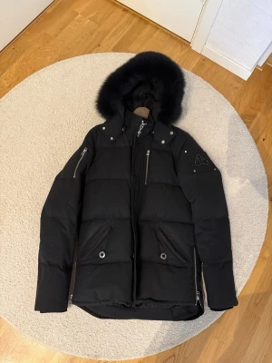 Moose Knuckles jacka 3Q - Säljer en svart dunjacka från Moose Knuckles med pälskantad huva och flera dragkedjor. Jackan har två stora fickor framtill, detaljerade metalldragkedjor och logga på ärmen. Perfekt för kalla vinterdagar och riktigt snygg streetstil.