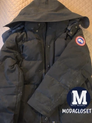 Svart dunjacka från Canada Goose - Svart dunjacka från Canada Goose med huva och klassisk logga på ärmen. Jackan har quiltad design, dragkedja framtill och är perfekt för kalla vinterdagar. Snygg och funktionell modell som håller dig varm hela säsongen. Lite smuts på armen men rengörs såklart innan köp!
