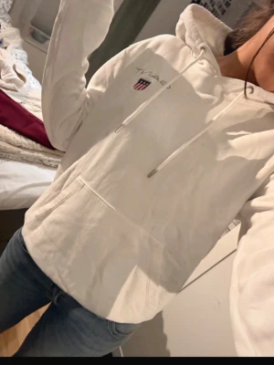 Vit hoodie från GANT med logga - Snygg vit hoodie från GANT med broderad logga och sköld på bröstet. Klassisk känguruficka framtill, dragsko i huvan och ribbade muddar. Perfekt för en chill och stilren look. Mjuk bomullsblandning som är skön att ha på sig.