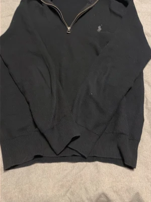 Svart halvzip tröja från Ralph Lauren - Klassisk svart tröja från Ralph Lauren med halvzip och diskret broderad logga på bröstet. Tröjan har ribbade muddar vid ärmslut och nederkant samt en mjuk, stickad känsla. Perfekt till jeans eller chinos för en clean look.