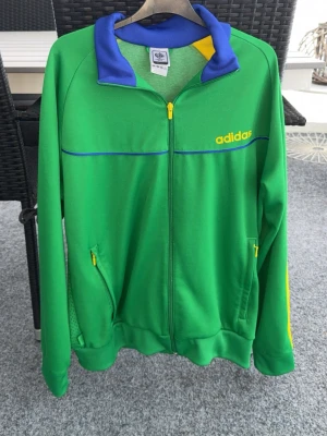 Grön Adidas Brasil träningsjacka - Snygg grön träningsjacka från Adidas med gula och blå detaljer samt gul dragkedja. Jackan har två fickor med dragkedja och texten 'adidas' på bröstet. På ryggen står det 'BRASIL' i stort tryck. Perfekt för sport eller streetwear.