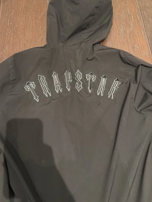Svart vindjacka från Trapstar - Säljer en svart vindjacka från Trapstar med stor logga broderad på ryggen och ikonisk T-dragkedja. Jackan har huva med snörning och en patch på ärmen med texten 'It's a secret'. Perfekt för dig som vill ha streetwear-vibe.
