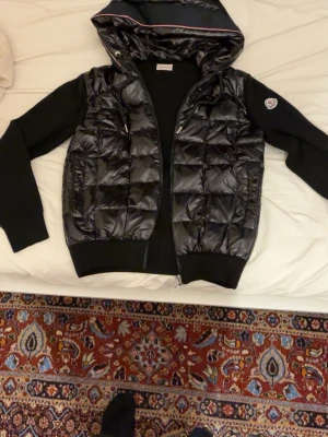 Svart dunjacka med huva från Moncler - Svart cardigan från Moncler med glansig quiltad front och huva. Jackan har långa ärmar i mjukt material, dragkedja framtill och en Moncler-logga på ärmen. Perfekt för dig som vill ha en stilren och sportig look.