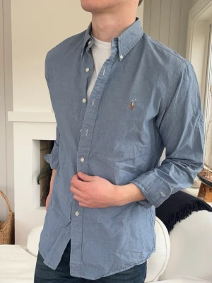 Blå skjorta från Polo Ralph Lauren - Klassisk blå skjorta från Polo Ralph Lauren med button-down krage och broderad logga på bröstet. Skjortan har lång ärm, knappar framtill och är tillverkad i bomull. Perfekt för en stilren och avslappnad look.