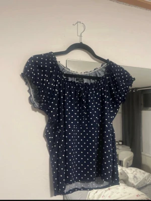 Blå prickig topp med volangärm - Mörkblå topp med vita prickar och bred resår vid halsen. Toppen har korta volangärmar och en lös, luftig passform. Perfekt för en trendig och avslappnad stil. Materialet känns lätt och svalt, troligen polyester eller bomullsblandning.  ‼️står 40-42 men sitter som M‼️
