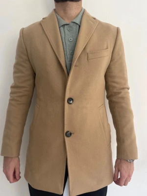 JL Beige ullkappa med knappar - Stilren beige kappa i ull med klassisk krage och två knappar framtill. Kappan har fickor på sidorna och en bröstficka för extra detalj. Perfekt för dig som gillar en clean och tidlös look.