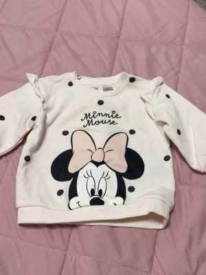 Vit sweatshirt med Minnie Mouse-tryck - Söt vit sweatshirt med svarta prickar och stort tryck av Minnie Mouse framtill. Tröjan har volangdetaljer vid axlarna och en ljusrosa rosett på Minnie. Rund halsringning och långa ärmar. Perfekt för dig som gillar Disney och vill ha en lekfull stil.