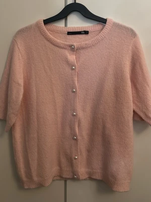Ljusrosa kofta med pärlknappar - Supersöt ljusrosa kofta från Lager 157 med korta ärmar och rund halsringning. Koftan har pärlknappar framtill och är gjord i ett mjukt, fluffigt material som känns riktigt mysigt. Perfekt för att addera en gullig touch till din outfit.