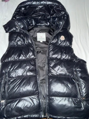 Svart dunväst från Moncler med huva - Snygg svart dunväst från Moncler med glansig finish och avtagbar huva. Västen har två dragkedjefickor framtill och klassisk Moncler-logga på bröstet. Perfekt för lager på lager och ger en sportig vibe till din outfit. Kan diskutera pris.