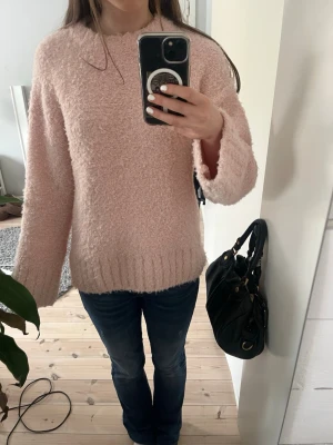 Ljusrosa fluffig stickad tröja - Mysig ljusrosa stickad tröja med fluffig textur och avslappnad passform. Tröjan har rund halsringning, långa ärmar och bred ribbstickad kant nedtill.