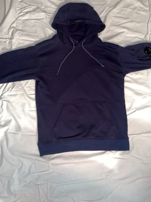 Mörkblå hoodie från C.P. Company - Snygg mörkblå hoodie från C.P. Company med klassisk huva och dragsnören. Hoodien har en stor magficka och en ikonisk linsdetalj på ena ärmen. Tillverkad i mjuk bomull för skön känsla och stilren look.