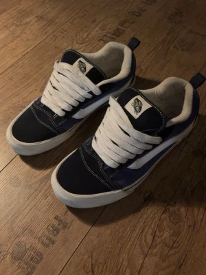 Marinblå och vita Vans sneakers - Säljer ett par klassiska Vans sneakers i marinblått och vitt med vit sula och breda vita snören. Skorna har den ikoniska sidostripen och rund tå. Ovandelen är i mocka och textil, perfekt för en avslappnad streetstil.