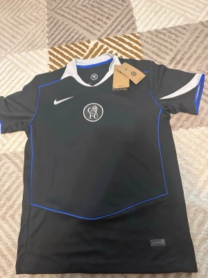 Chelsea FC svart matchtröja Nike - Snygg svart Chelsea FC matchtröja från Nike med blå detaljer och vit krage. Tröjan har klubbmärke på bröstet och är tillverkad i Dri-FIT material som andas. Perfekt för fotbollsträning eller att bära till vardags.