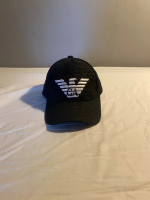 Svart keps från Armani med logga - Snygg svart keps från Armani med stor vit broderad logga framtill och diskret mönster med texten 'Giorgio Armani' över hela kepsen. Klassisk böjd skärm och justerbar baksida. Perfekt för dig som vill ha en stilren accessoar med lyxig känsla.